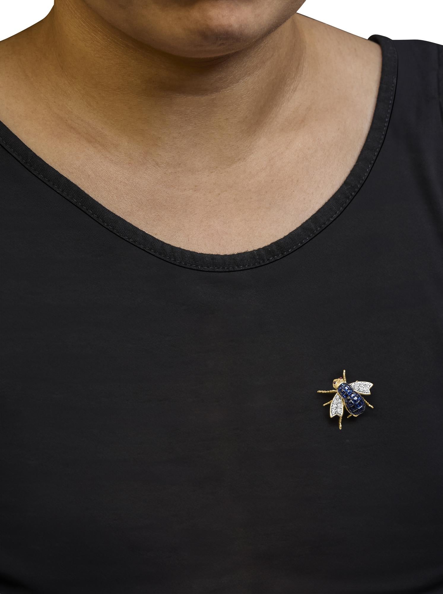 Yellow metal bee-shaped brooch with blue cushion-shaped gemstones on the body, white round pave-set gemstones on the wings, and two red round bezel-set gemstones on the head, shown on a model from a front angle.