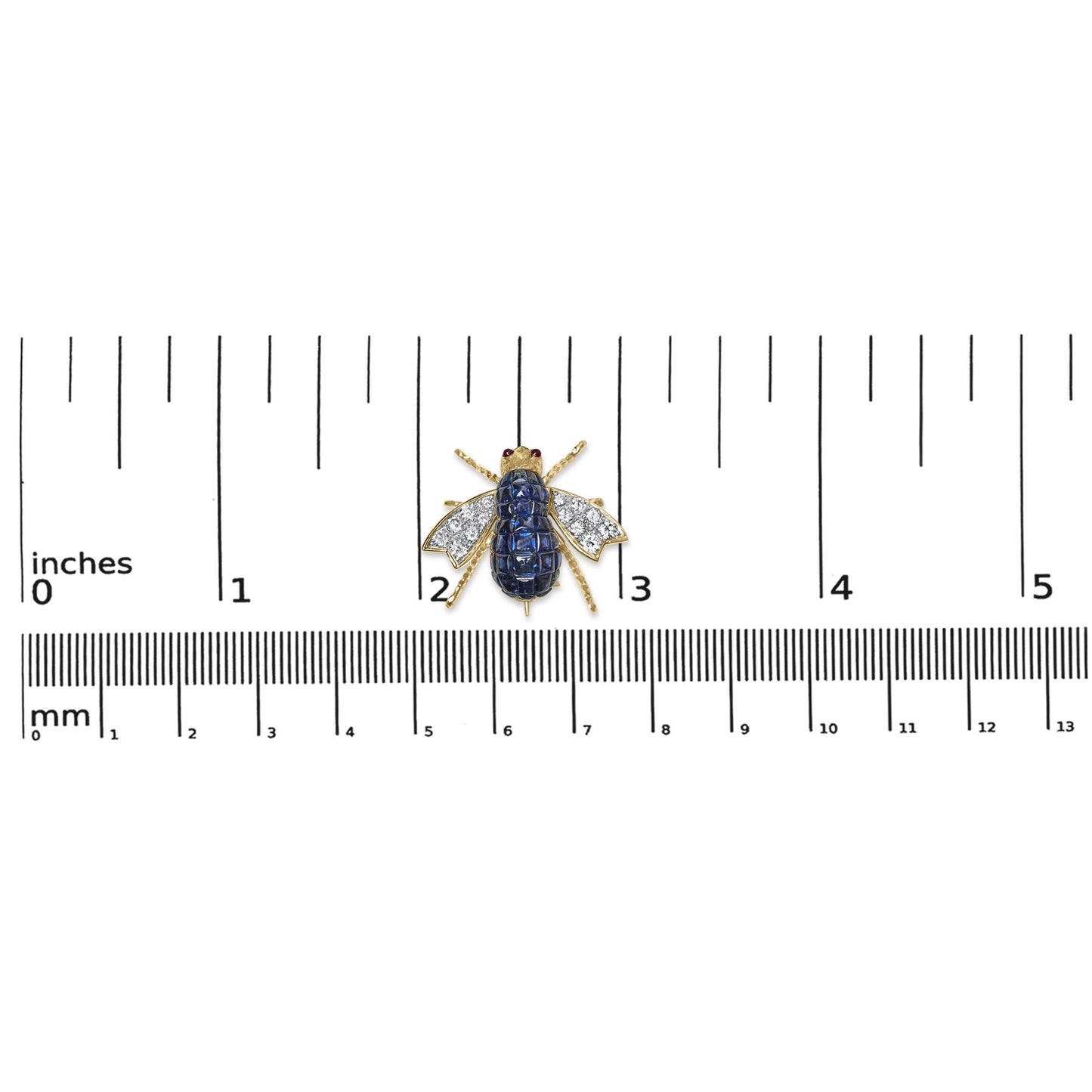 Yellow metal bee-shaped brooch with blue cushion-shaped gemstones on the body, white round pave-set gemstones on the wings, and two red round bezel-set gemstones for eyes, photographed from above on a ruler for size reference.