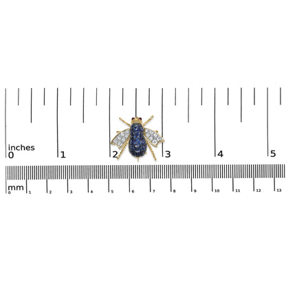 Yellow metal bee-shaped brooch with blue cushion-shaped gemstones on the body, white round pave-set gemstones on the wings, and two red round bezel-set gemstones for eyes, photographed from above on a ruler for size reference.