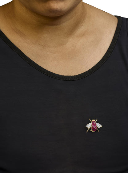 Close-up front view of a yellow metal insect-shaped brooch worn on a model's black top, featuring red cushion-shaped gemstones on the body, white round pave-set gemstones on the wings, and two green round bezel-set gemstones on the head area.