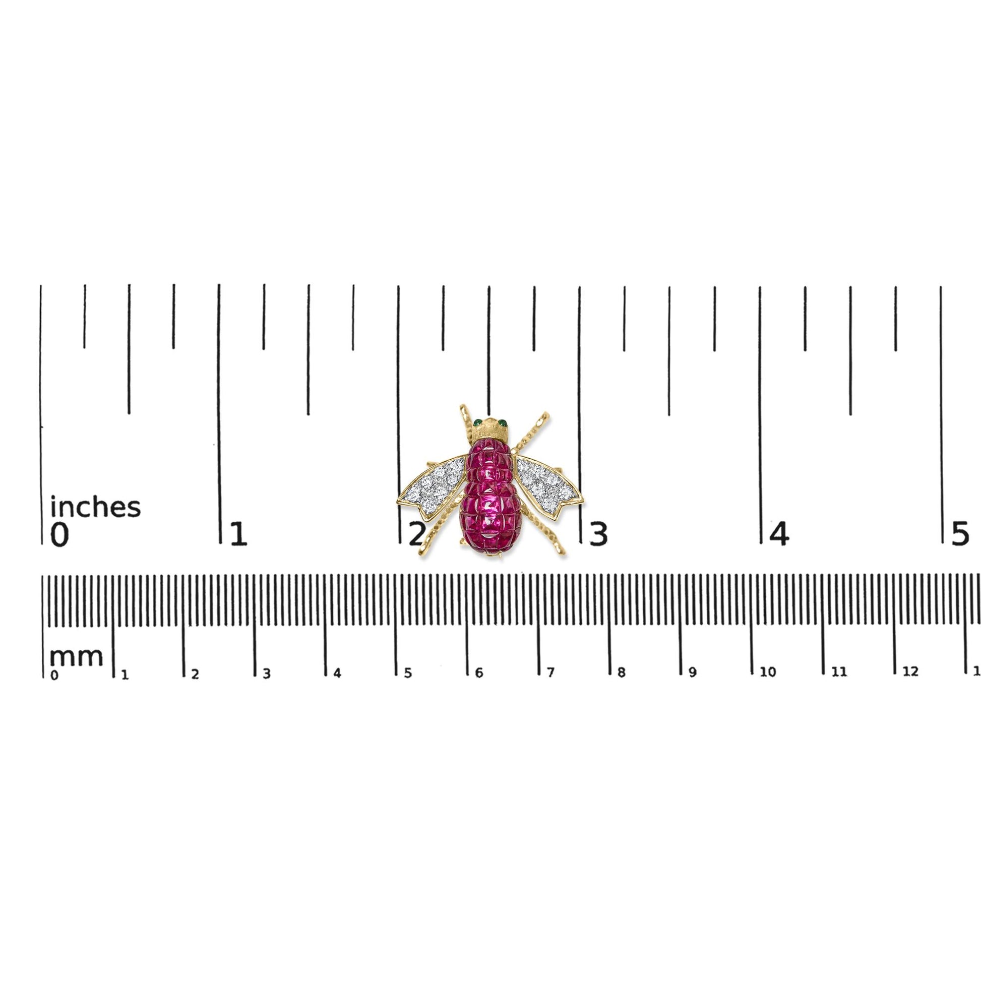 Top-down view of a yellow metal insect-shaped brooch with red gemstones on the body, white gemstones on the wings, and green gemstones on the eyes, placed on a ruler for size reference.