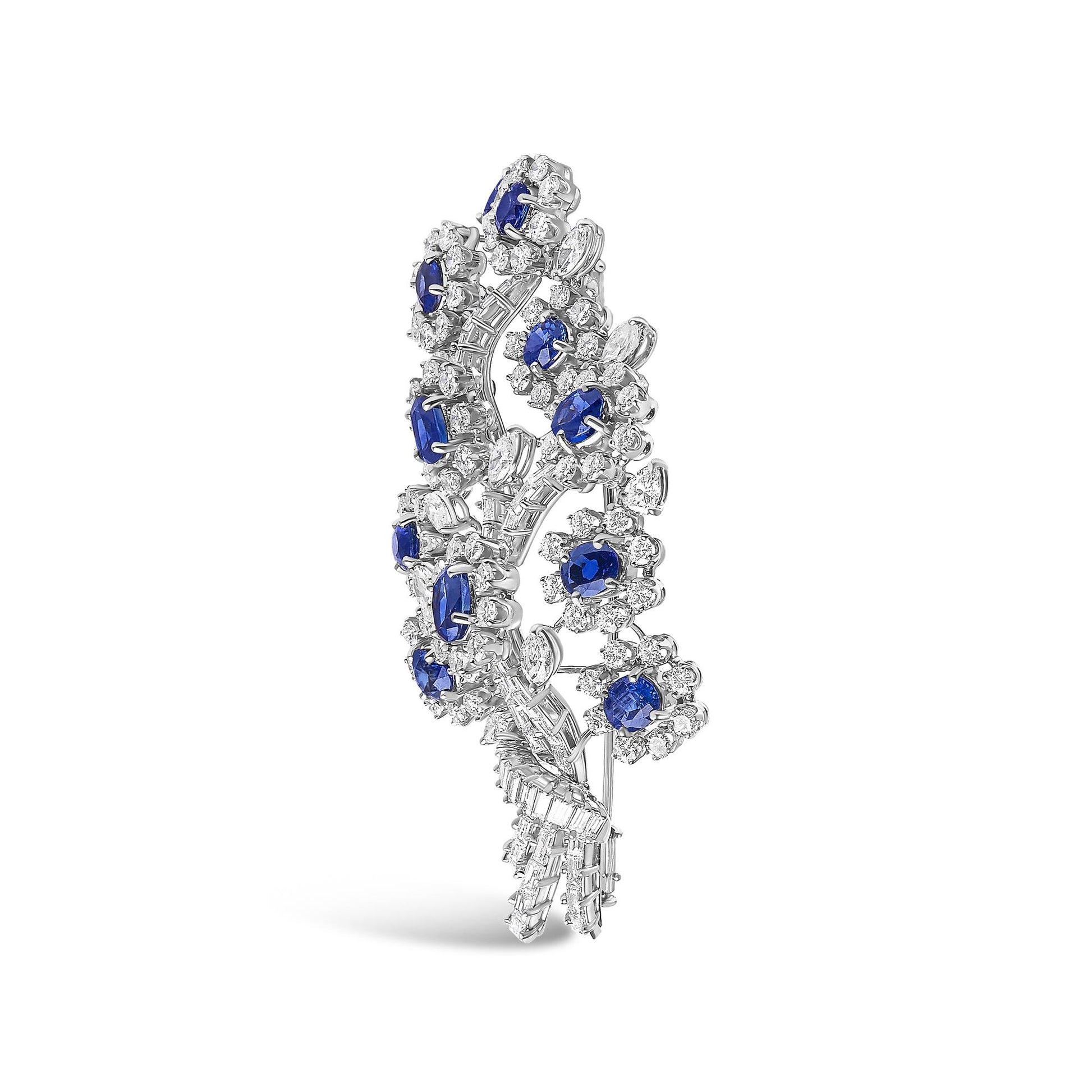 White-colored brooch with a vertical, curved floral design featuring clustered white gemstones in round, marquise, pear, and baguette shapes surrounding oval blue gemstones, shown at a slight angle on a white background.