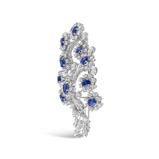 White-colored brooch with a vertical, curved floral design featuring clustered white gemstones in round, marquise, pear, and baguette shapes surrounding oval blue gemstones, shown at a slight angle on a white background.