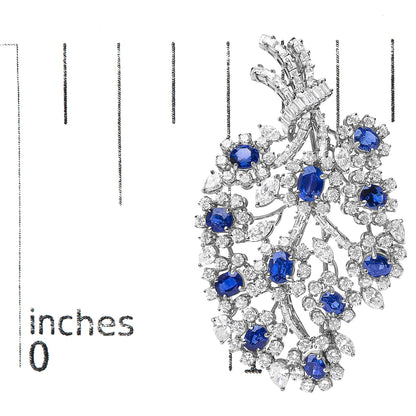 White metal brooch shaped like a floral cluster with oval blue gemstones surrounded by variously shaped white gemstones, shown on a scale from a front angled view.