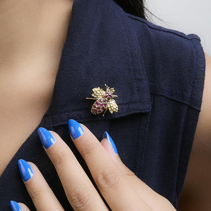 Close-up angled shot of a yellow metal bee-shaped brooch with round red gemstones on the body, worn on the collar of a dark textured fabric by a model with blue manicured nails.