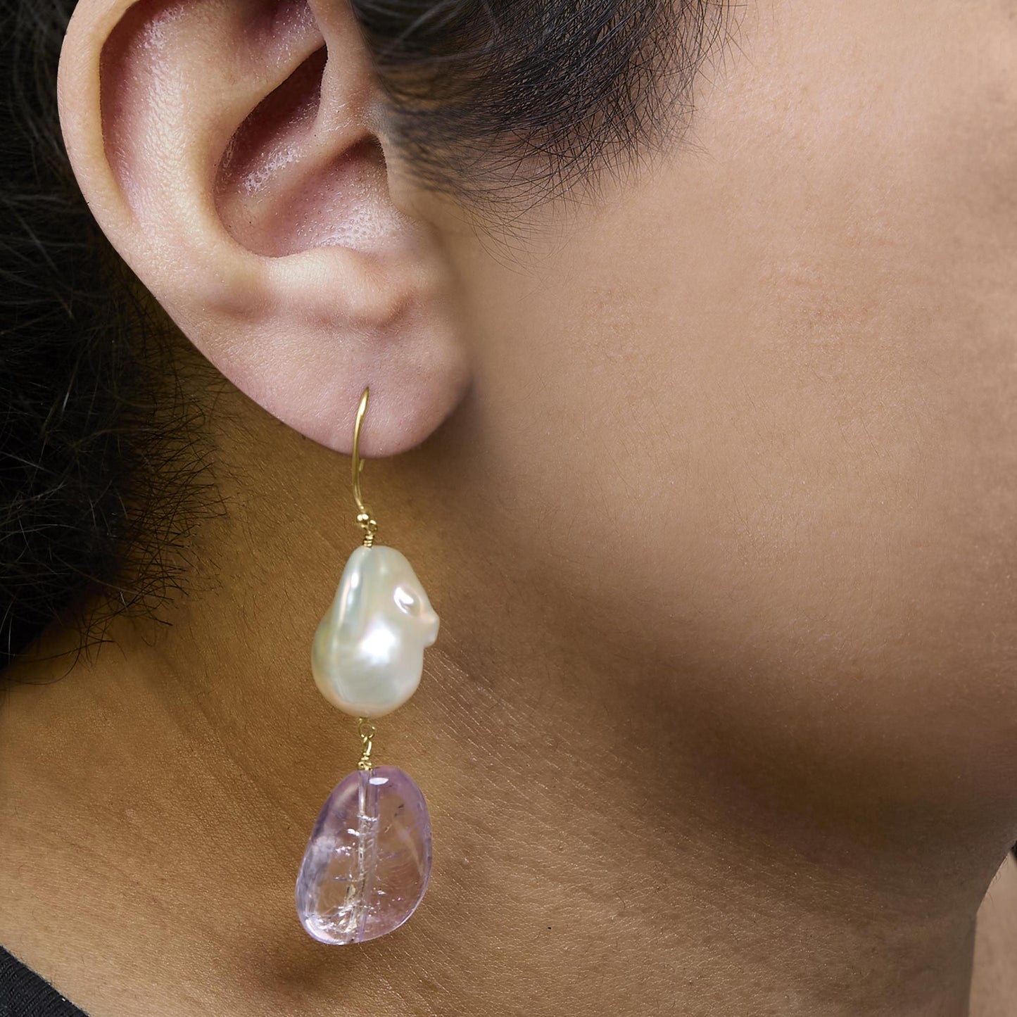 Close-up side view of a model wearing a yellow metal earring featuring an irregular white gemstone above a purple gemstone in drilled settings.