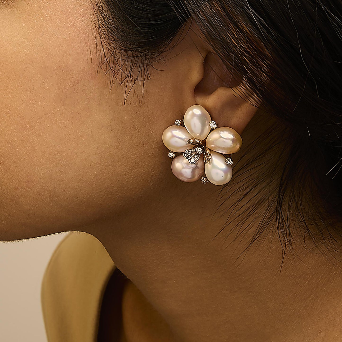 Rose metal earrings shaped like a flower with five large pearl-like petals and small white and champagne gemstones, shown worn on a model from a side angle.
