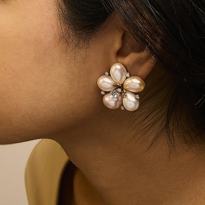 Rose metal earrings shaped like a flower with five large pearl-like petals and small white and champagne gemstones, shown worn on a model from a side angle.