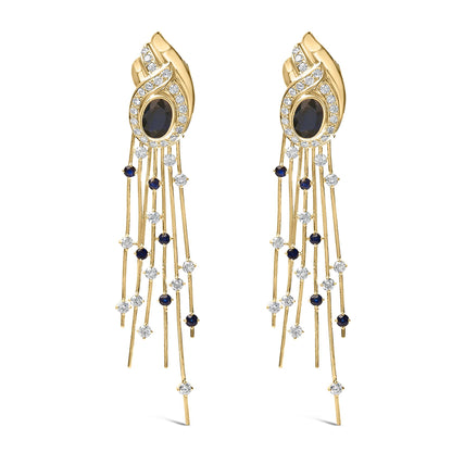 Pair of yellow metal earrings shown from the front, featuring an oval blue gemstone in a bezel setting surrounded by a swirl of white gemstones in a pave setting, with multiple thin strands dangling below adorned with round blue and white gemstones in prong settings.