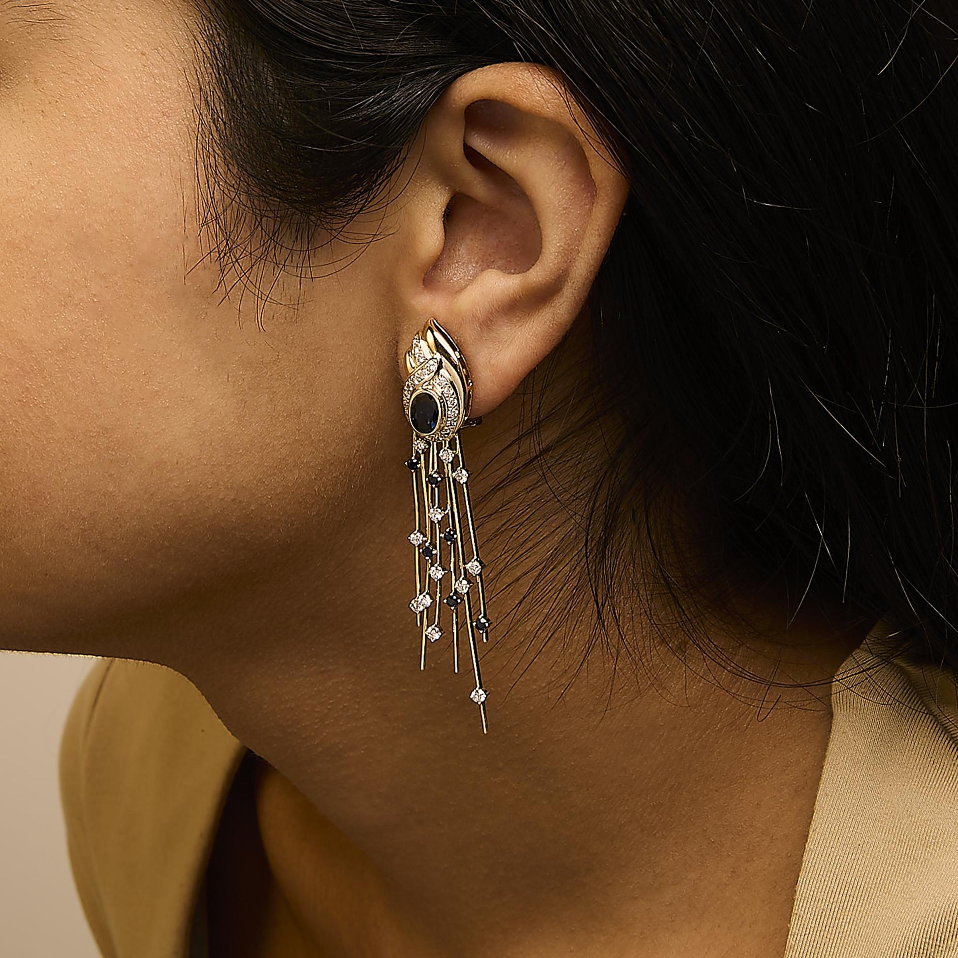 Yellow metal long dangle earrings with an oval blue gemstone in bezel setting, surrounded by white gemstones in pave and prong settings, worn on a model, side angle view.