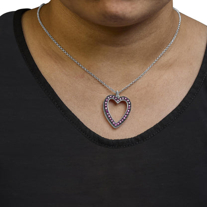 Close-up front view of a white metal heart-shaped necklace pendant with small round red gemstones set around the edges and a few small round white gemstones, worn on a model.