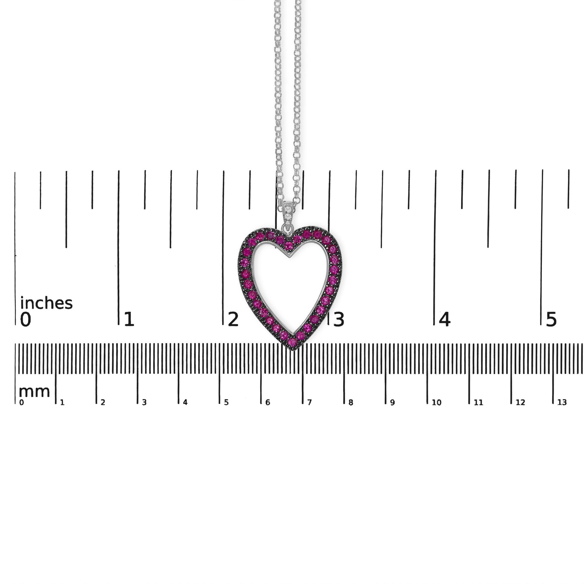 White metal heart-shaped necklace pendant with round red gemstones along the border and three small round white gemstones at the top, shown hanging against a ruler for size reference, front-facing angle.