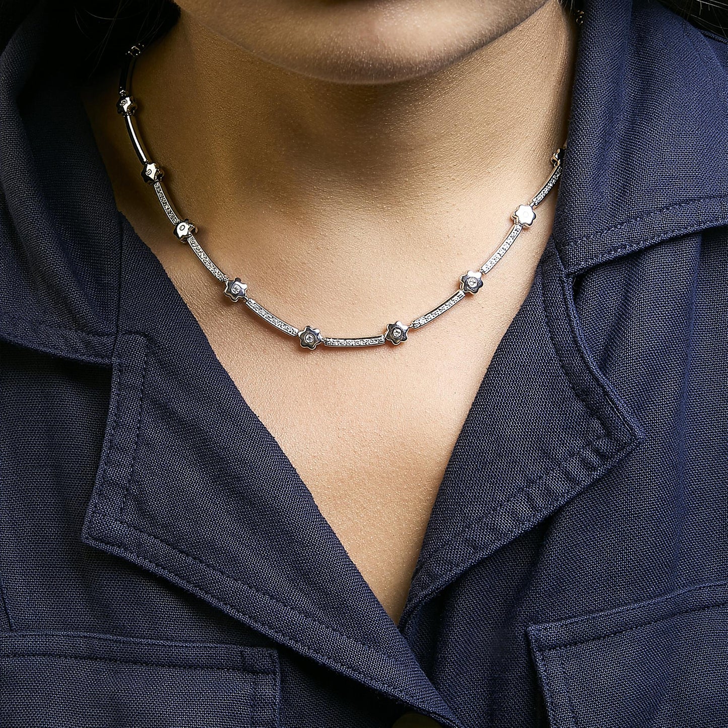White metal necklace with alternating curved bars and small flower-shaped links, each set with white round gemstones, shown worn on a model from a front angle.
