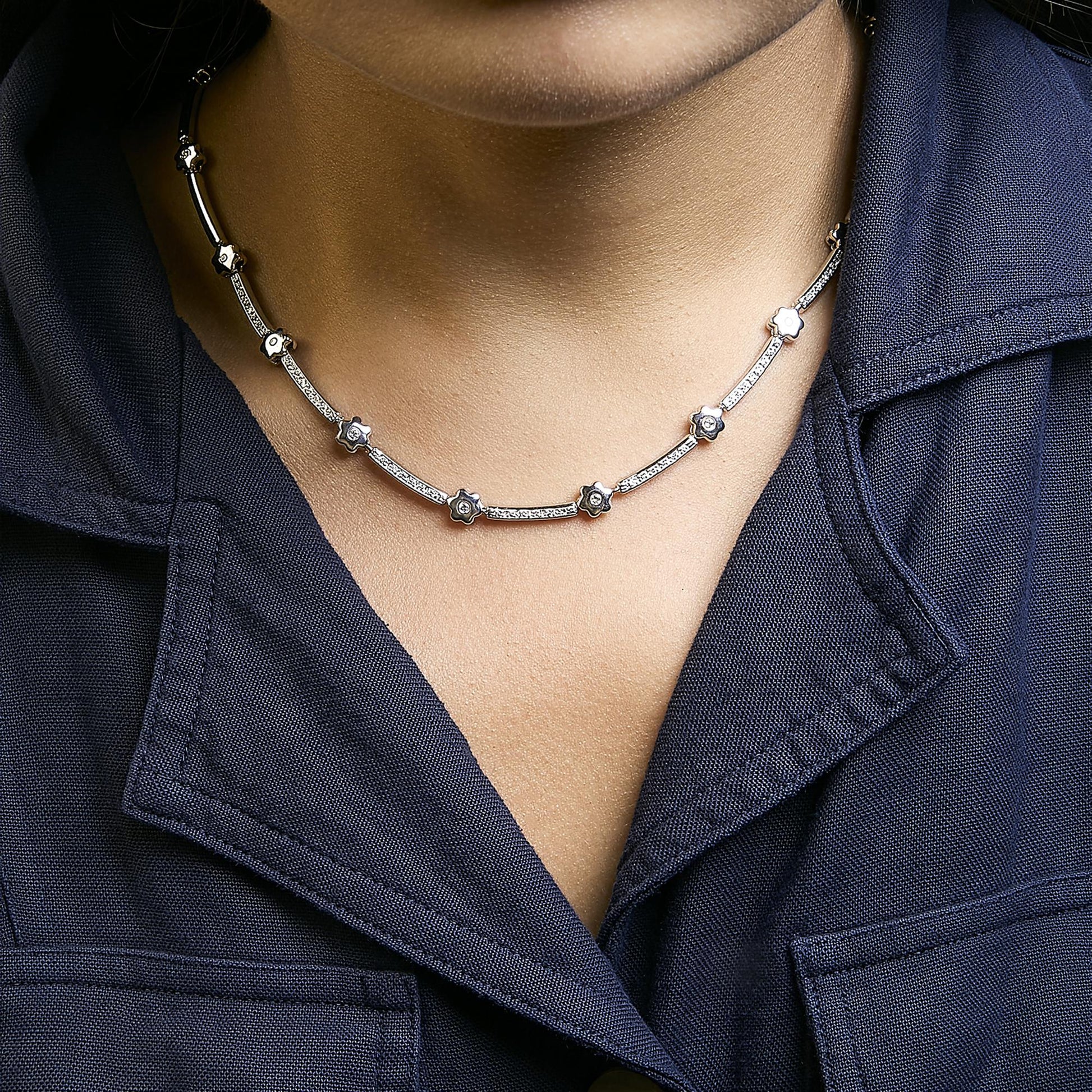White metal necklace with alternating curved bars and small flower-shaped links, each set with white round gemstones, shown worn on a model from a front angle.