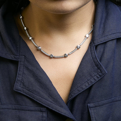 White metal necklace with alternating curved bars and small flower-shaped links, each set with white round gemstones, shown worn on a model from a front angle.