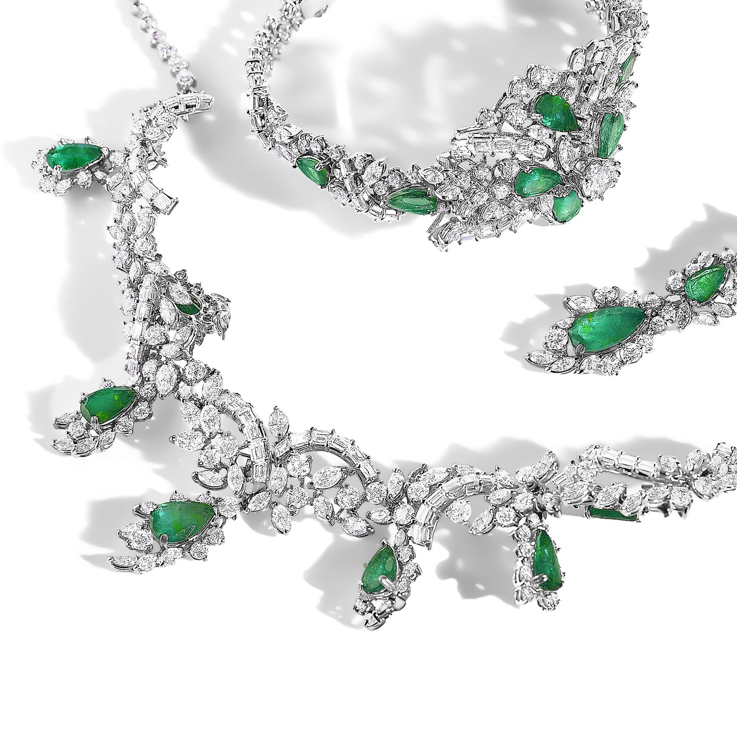 Set of jewelry pieces displayed in a flat lay, featuring white metal adorned with pear-shaped green gemstones and variously shaped white gemstones, including round, marquise, and baguette cuts, arranged in intricate patterns.