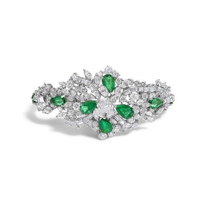 Top-down angle of a white metal bracelet set with pear-shaped green gemstones and a variety of round, baguette, and marquise-shaped white gemstones arranged in an intricate floral-inspired design.