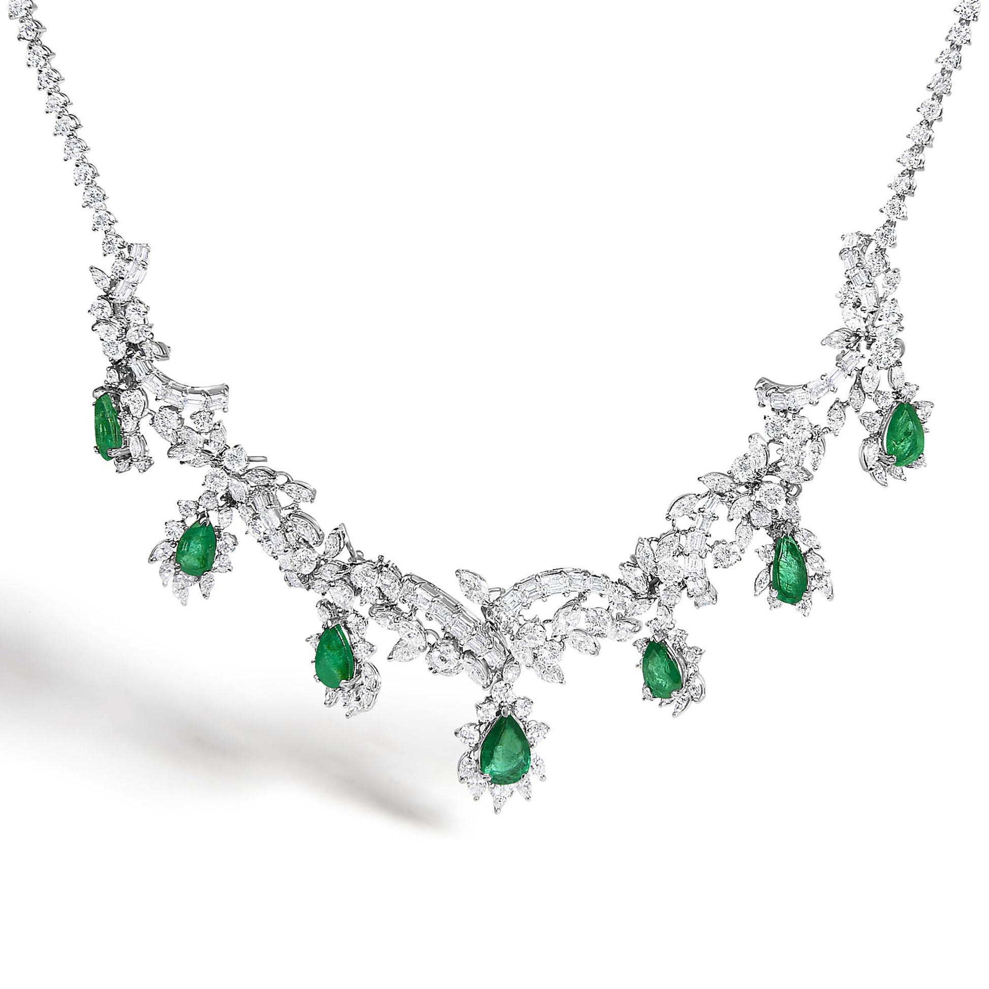 Close-up front angle of a white metal necklace set featuring intricate design with round, baguette, and marquise shaped white gemstones, accented with pear-shaped green gemstones evenly spaced along the piece.