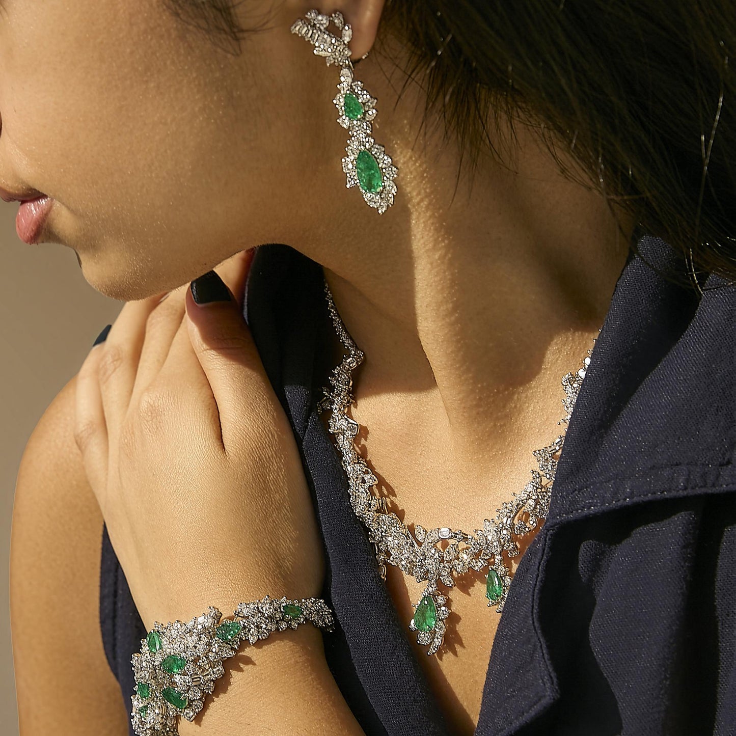 Set of jewelry shown worn on a model featuring long earrings with green pear-shaped gemstones surrounded by white gemstones, a matching necklace with green pear-shaped gemstone pendants and intricate white gemstone detailing, and a bracelet with similar green pear-shaped gemstones and white gemstone accents, photographed from a close-up side angle.