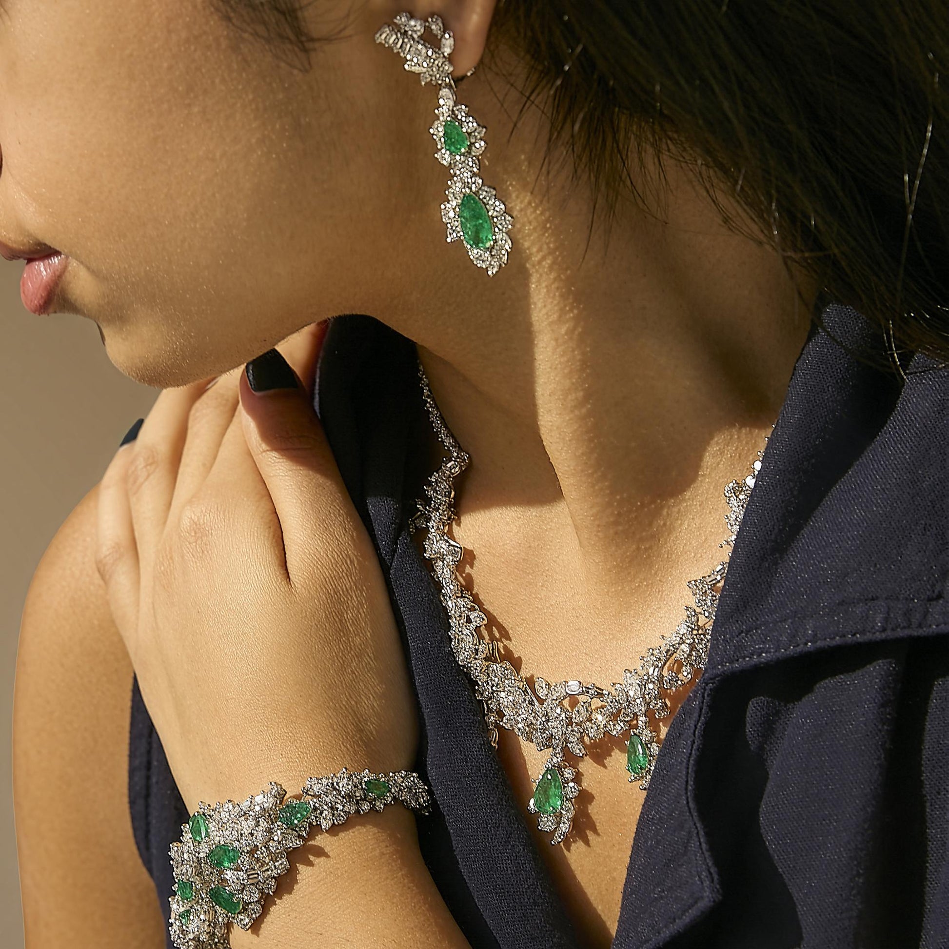 Set of jewelry shown worn on a model featuring long earrings with green pear-shaped gemstones surrounded by white gemstones, a matching necklace with green pear-shaped gemstone pendants and intricate white gemstone detailing, and a bracelet with similar green pear-shaped gemstones and white gemstone accents, photographed from a close-up side angle.