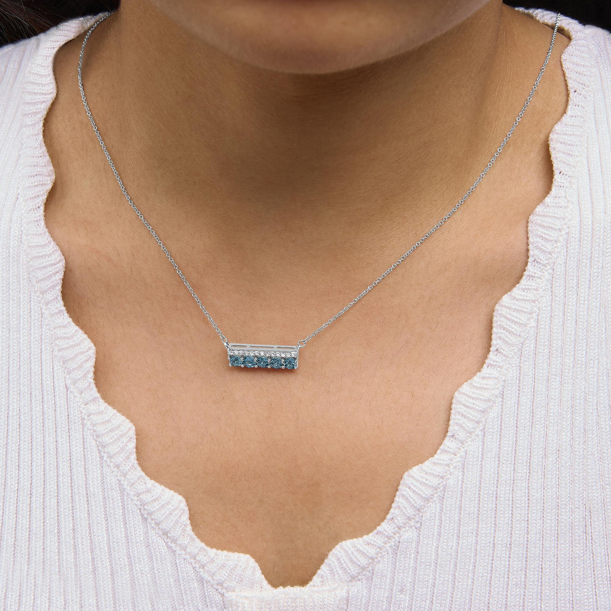 White metal necklace with a rectangular pendant featuring round blue gemstones in prong settings along the bottom edge and white gemstones in pave settings above, shown worn on a model from a front angle.