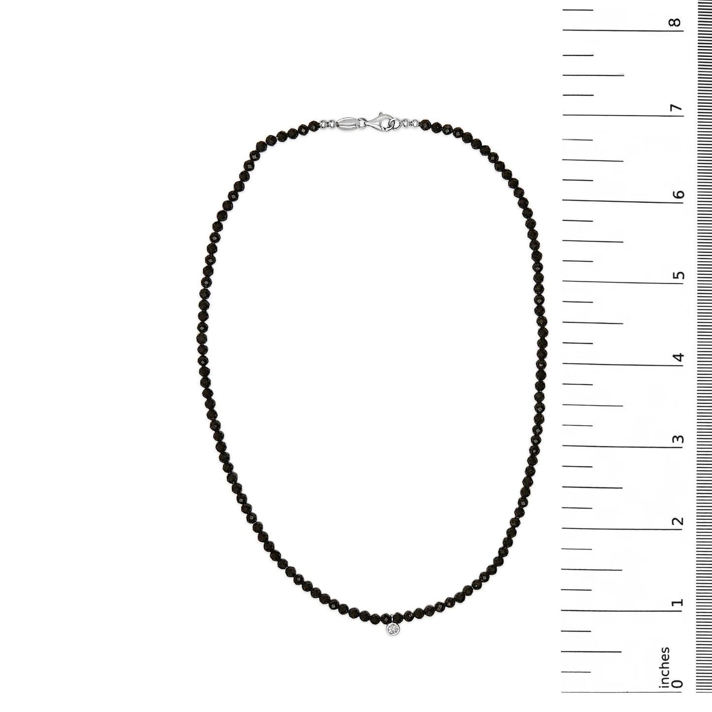 White metal necklace with small round black gemstones strung along its length and a single small round white gemstone pendant, shown on a flat surface with a vertical ruler for scale, top-down angle.