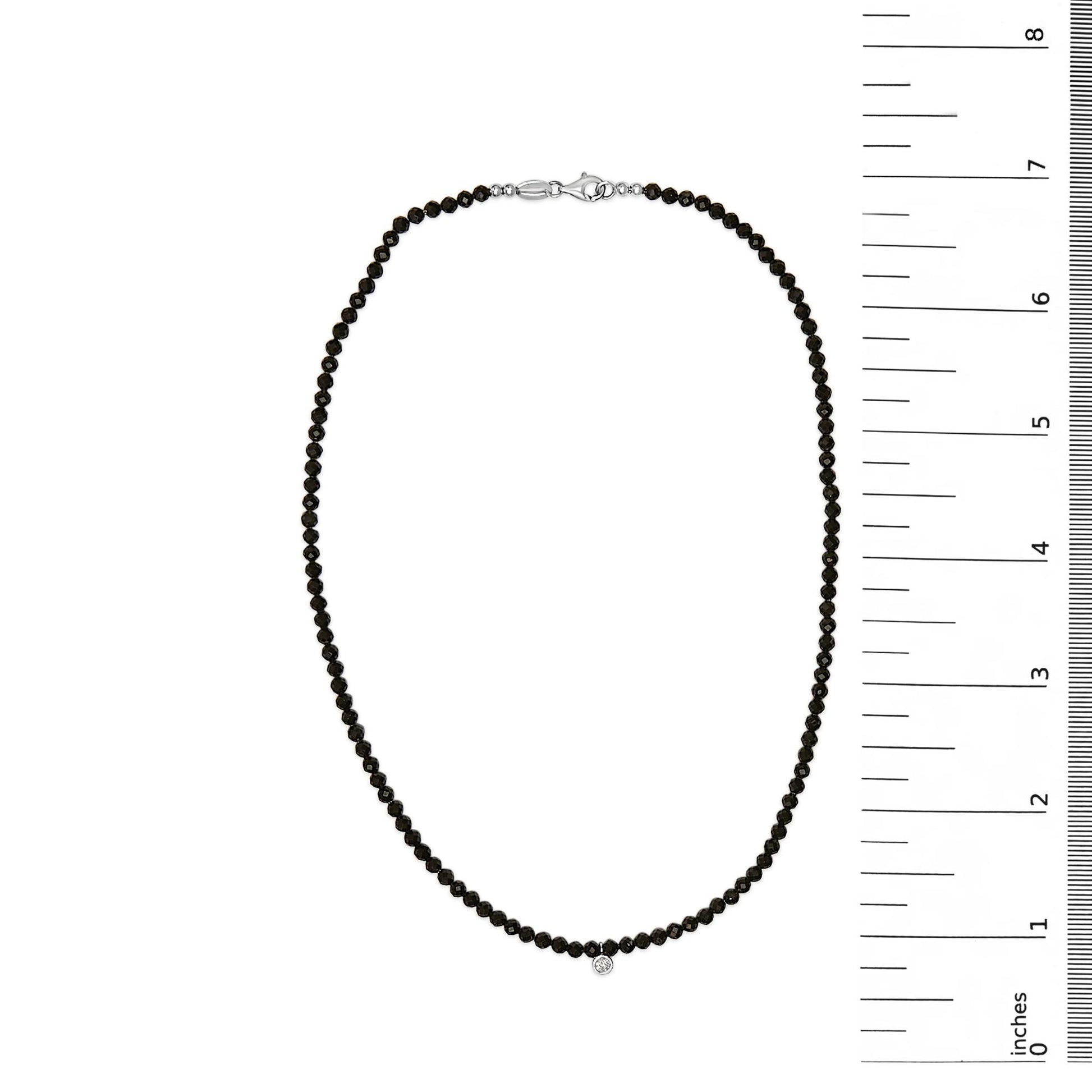 White metal necklace with small round black gemstones strung along its length and a single small round white gemstone pendant, shown on a flat surface with a vertical ruler for scale, top-down angle.