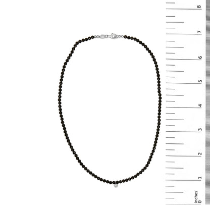 White metal necklace with small round black gemstones strung along its length and a single small round white gemstone pendant, shown on a flat surface with a vertical ruler for scale, top-down angle.