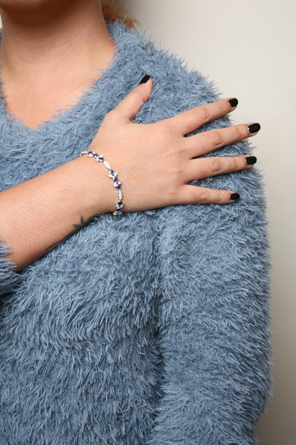 White metal bracelet with alternating round white gemstones in prong settings and round blue gemstones in bezel settings, shown on a model’s wrist from an angled top-down view.
