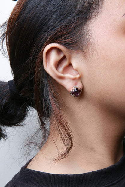 Close-up side view of a rose-colored earring on a model's ear, featuring a purple gemstone in an invisible setting surrounded by small white gemstones in prong settings.