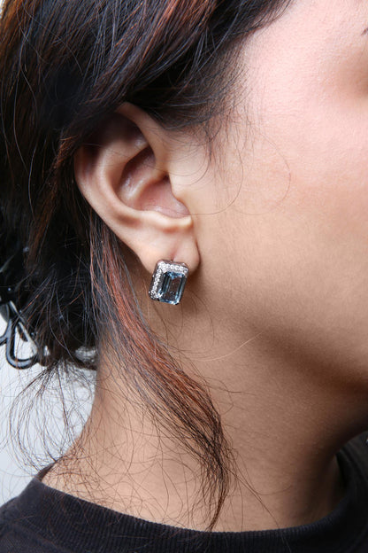 Close-up side view of a white metal earring on a model's ear, featuring a large blue emerald-shaped gemstone surrounded by small round white gemstones in a prong setting.