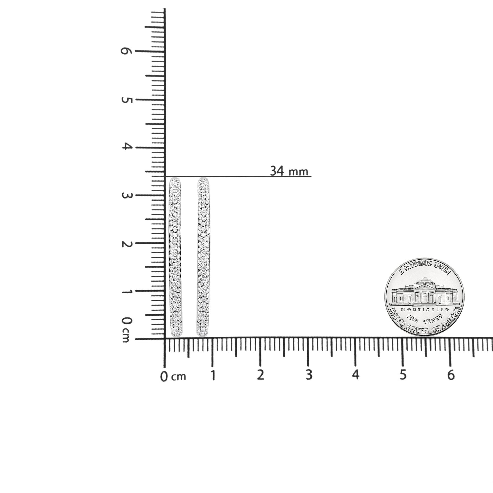 Close-up front view of white metal hoop earrings with rows of round white gemstones in prong settings, displayed on a ruler and next to a coin for size reference.