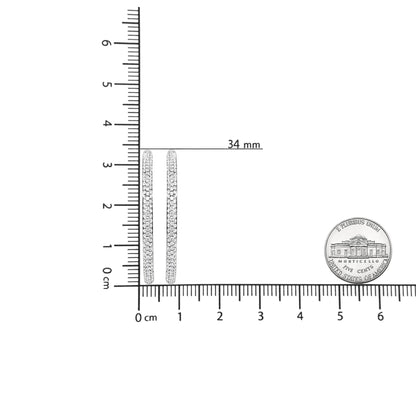 Close-up front view of white metal hoop earrings with rows of round white gemstones in prong settings, displayed on a ruler and next to a coin for size reference.