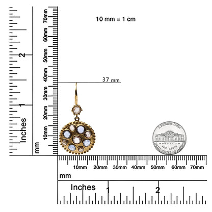Close-up front view of a yellow metal earring with round blue and brown bezel-set gemstones, displayed next to rulers and a coin for size reference.