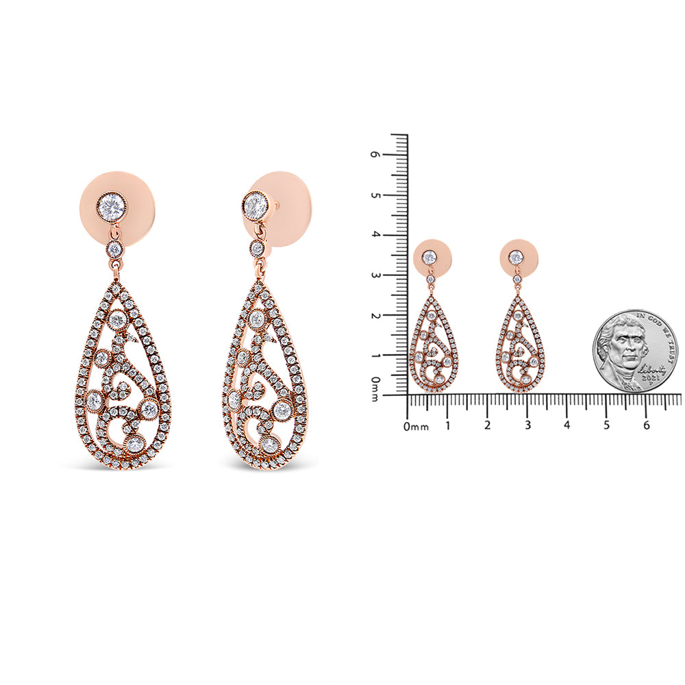 Front view of a pair of rose-colored metal earrings with a teardrop shape adorned with multiple small white gemstones in a scrolling pattern, shown next to a ruler and a coin for size reference.
