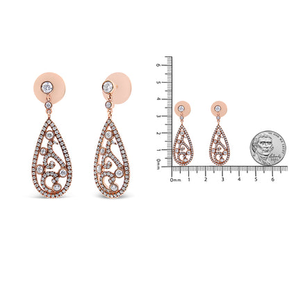 Front view of a pair of rose-colored metal earrings with a teardrop shape adorned with multiple small white gemstones in a scrolling pattern, shown next to a ruler and a coin for size reference.