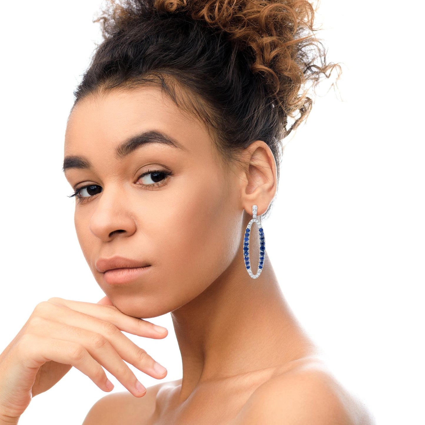 White metal oval-shaped earrings with white and blue gemstones in prong settings, worn on a model shown in a close-up side angle.