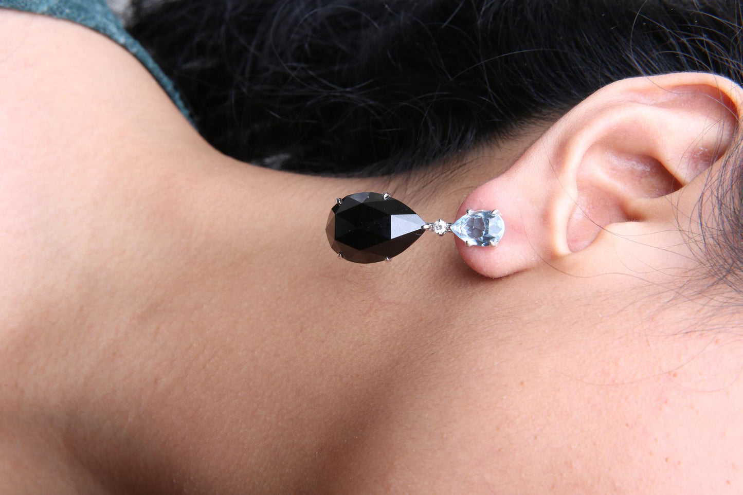 Close-up side view of a white metal earring on a model's ear, featuring a pear-shaped blue gemstone at the top, a small round white gemstone below it, and a larger pear-shaped black gemstone hanging beneath.