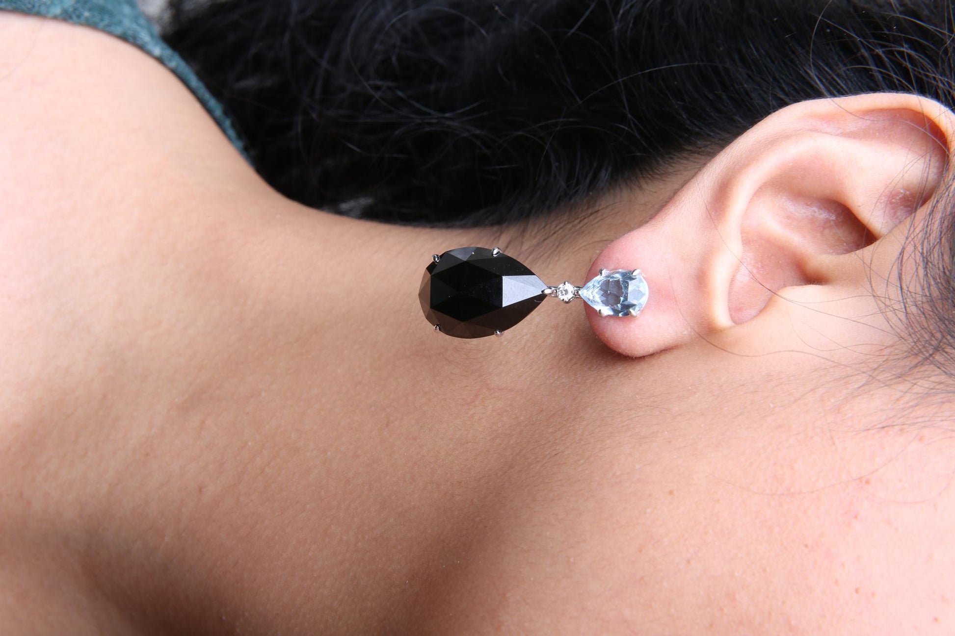 Close-up side view of a white metal earring on a model's ear, featuring a pear-shaped blue gemstone at the top, a small round white gemstone below it, and a larger pear-shaped black gemstone hanging beneath.