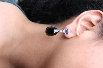 Close-up side view of a white metal earring on a model's ear, featuring a pear-shaped blue gemstone at the top, a small round white gemstone below it, and a larger pear-shaped black gemstone hanging beneath.