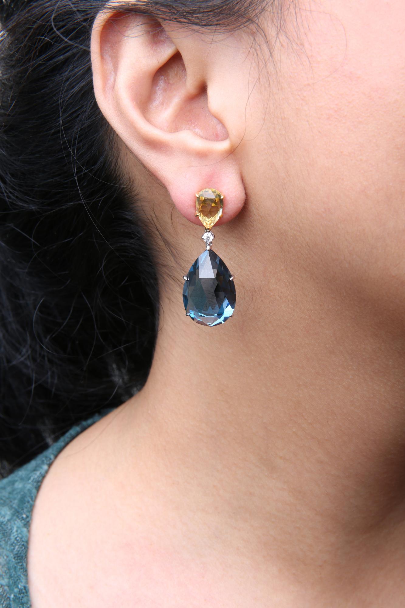 Close-up side view of an earring on a model featuring a yellow pear-shaped gemstone at the top, a small round white gemstone below it, and a larger blue pear-shaped gemstone dangling at the bottom, all set in yellow and white metal.