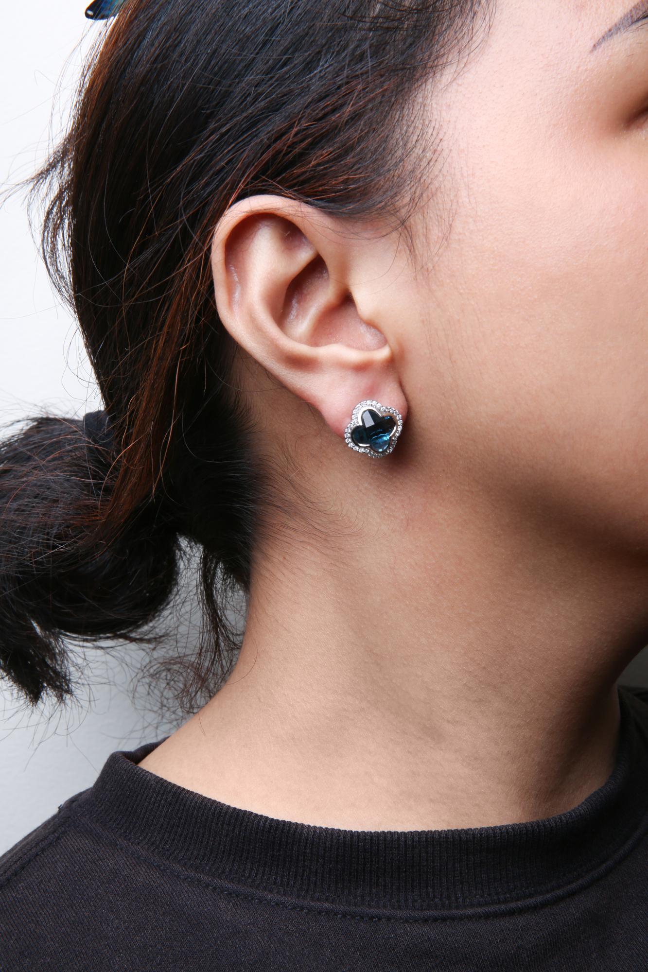 Close-up side view of a white metal earring on a model's ear, featuring a clover-shaped design with blue gemstones and surrounded by small white round gemstones in prong settings.