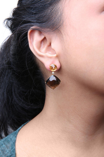 Close-up side view of a model wearing yellow metal earrings featuring a round yellow gemstone at the top, a small round white gemstone beneath it, and a cushion-shaped smoky gemstone dangling below.