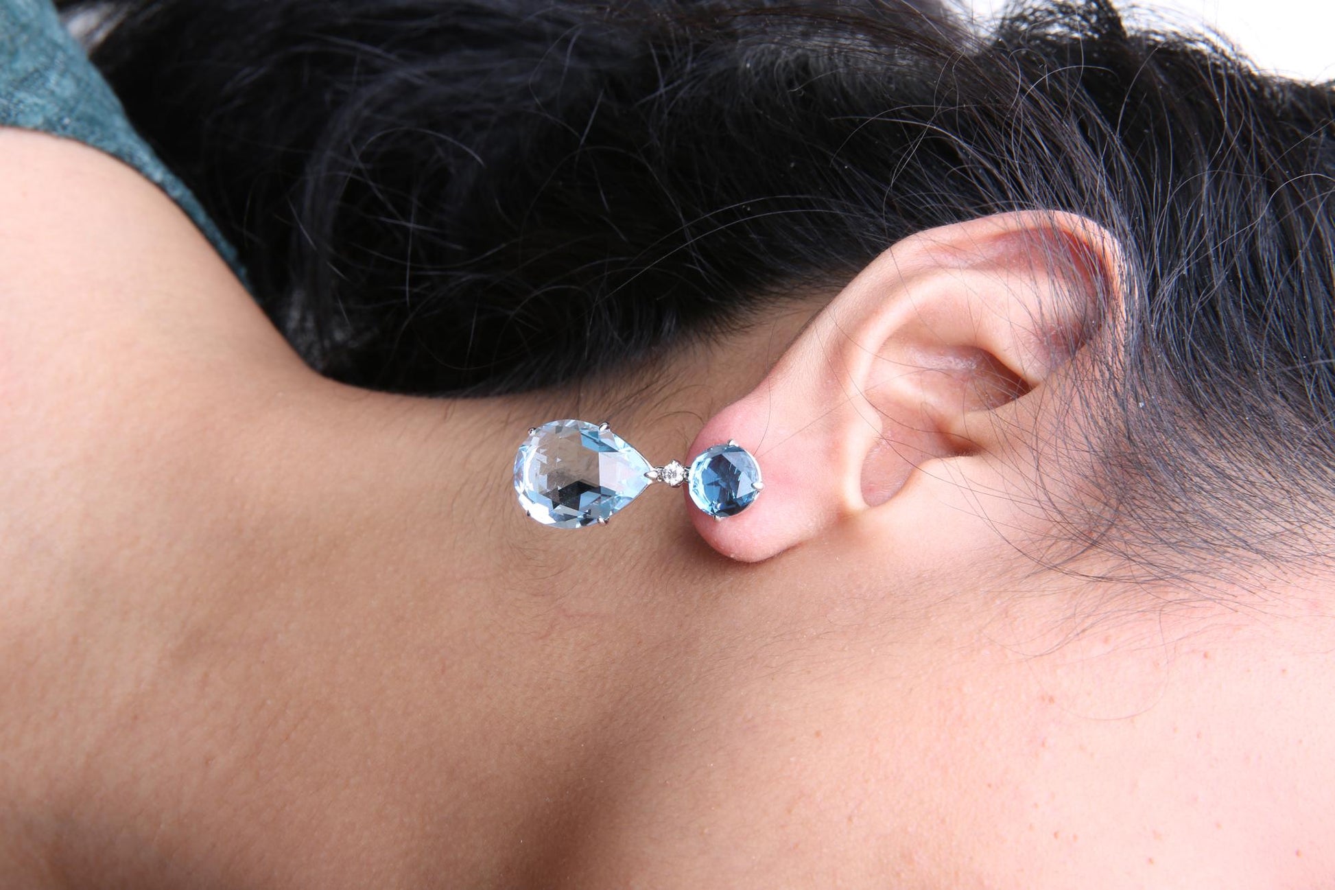 Close-up side view of a white metal earring on a model, featuring a round blue gemstone at the top, a small round white gemstone below it, and a larger pear-shaped blue gemstone dangling beneath.