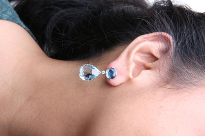 Close-up side view of a white metal earring on a model, featuring a round blue gemstone at the top, a small round white gemstone below it, and a larger pear-shaped blue gemstone dangling beneath.