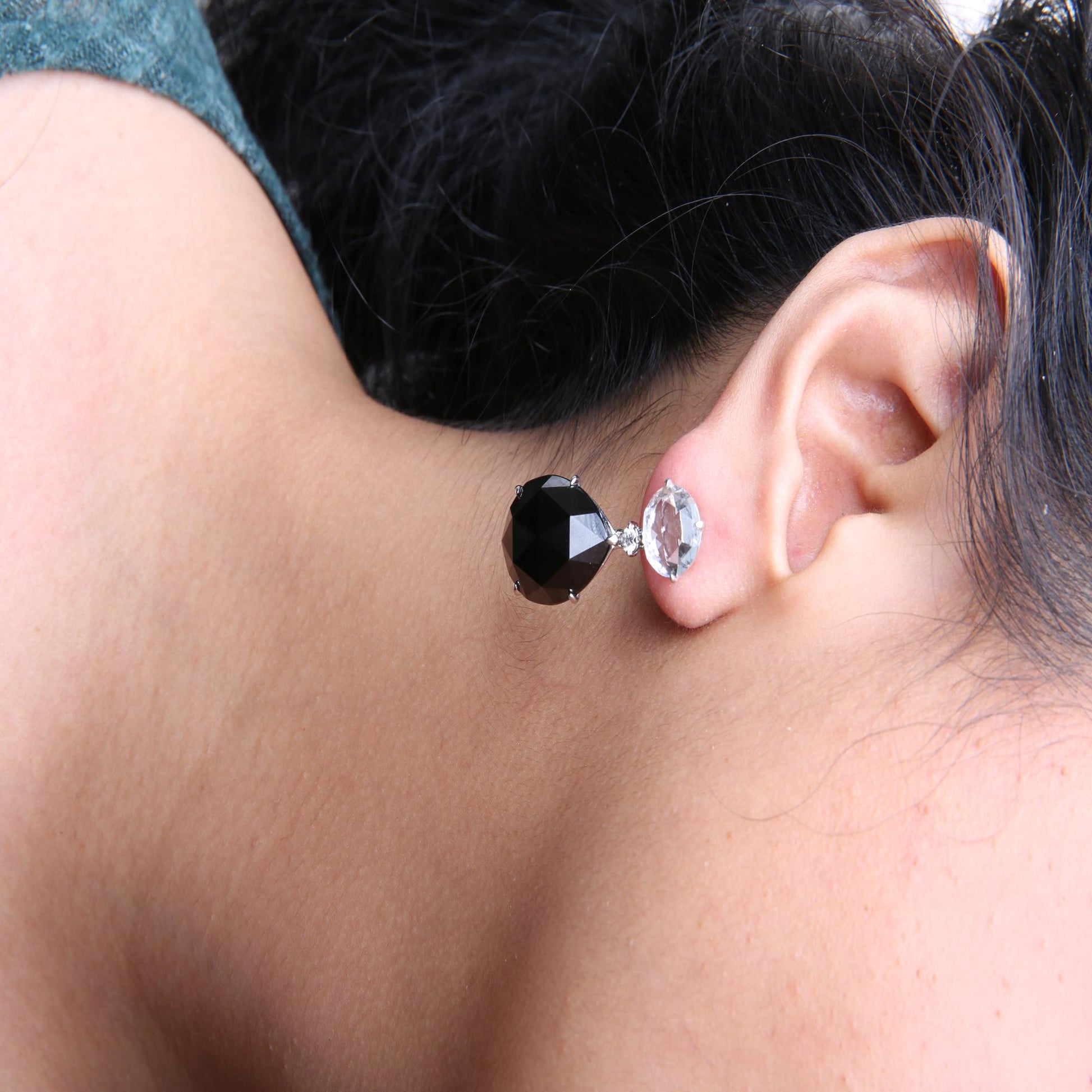 Close-up side view of a white metal earring worn on a model's ear, featuring a large round white gemstone at the top, a small round white gemstone below it, and a pear-shaped black gemstone dangling underneath.