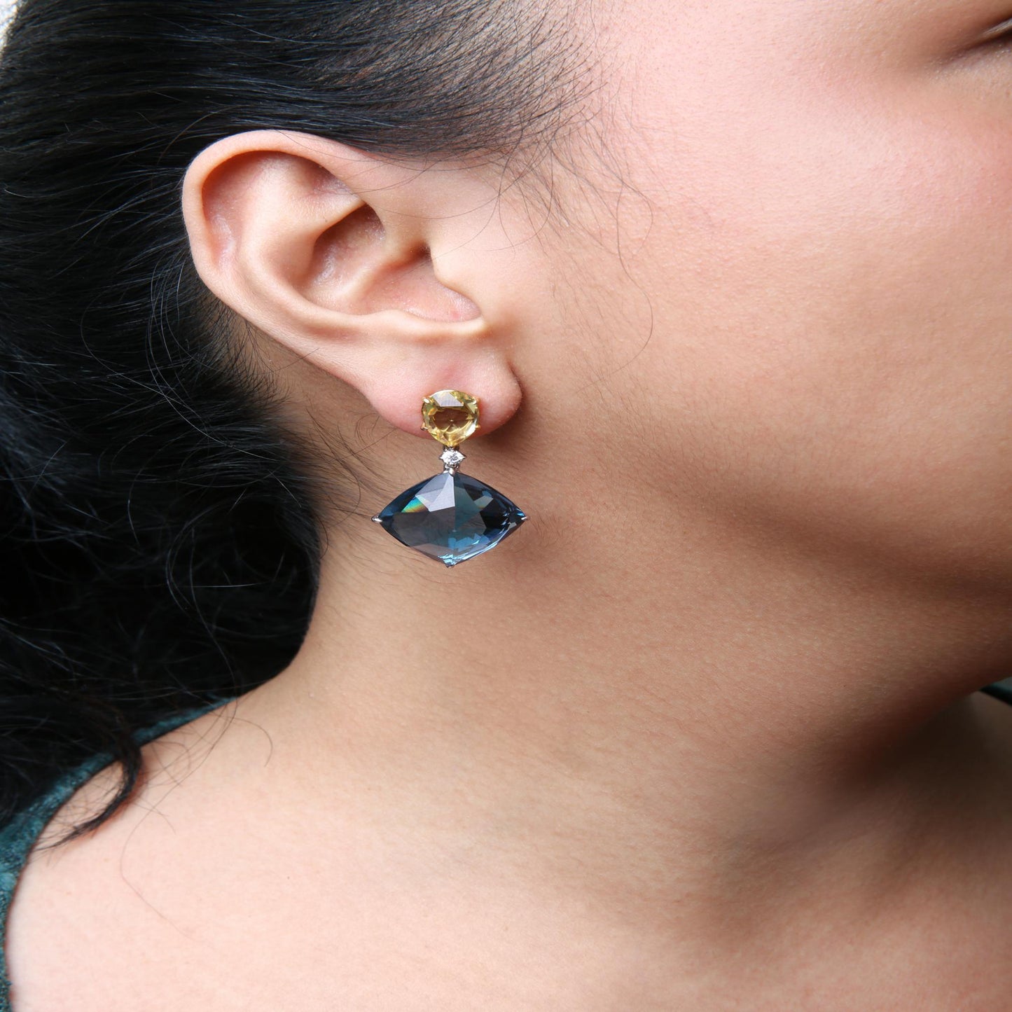 Close-up side view of a model wearing earrings featuring a yellow pear-shaped gemstone at the top, a small round white gemstone in the middle, and a large blue cushion-shaped gemstone dangling below in prong settings with yellow and white metal.
