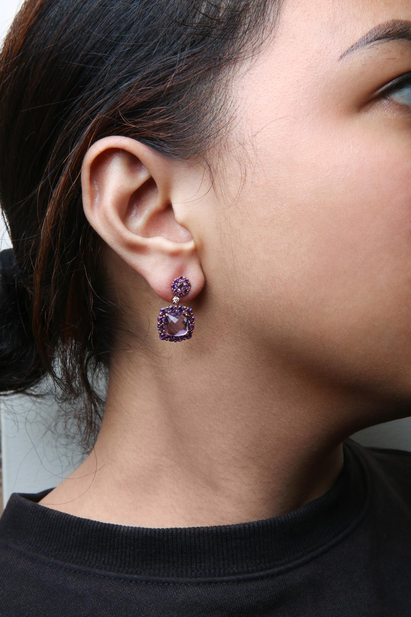 Close-up side angle of a model wearing rose metal earrings featuring a round cluster of purple gemstones at the stud, a small white round gemstone below, and a larger cushion-shaped purple gemstone surrounded by round purple gemstones dangling below.
