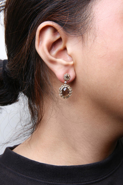Close-up side view of rose-colored metal drop earrings on a model, featuring oval smoky gemstones surrounded by smaller round lemon-colored and smoky gemstones, with a white gemstone connector.