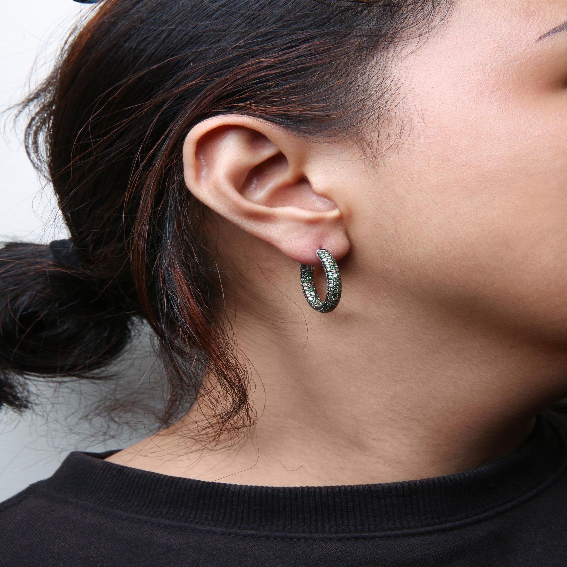 Close-up side view of a model wearing white metal hoop earrings set with round green and brown gemstones and white gemstones.